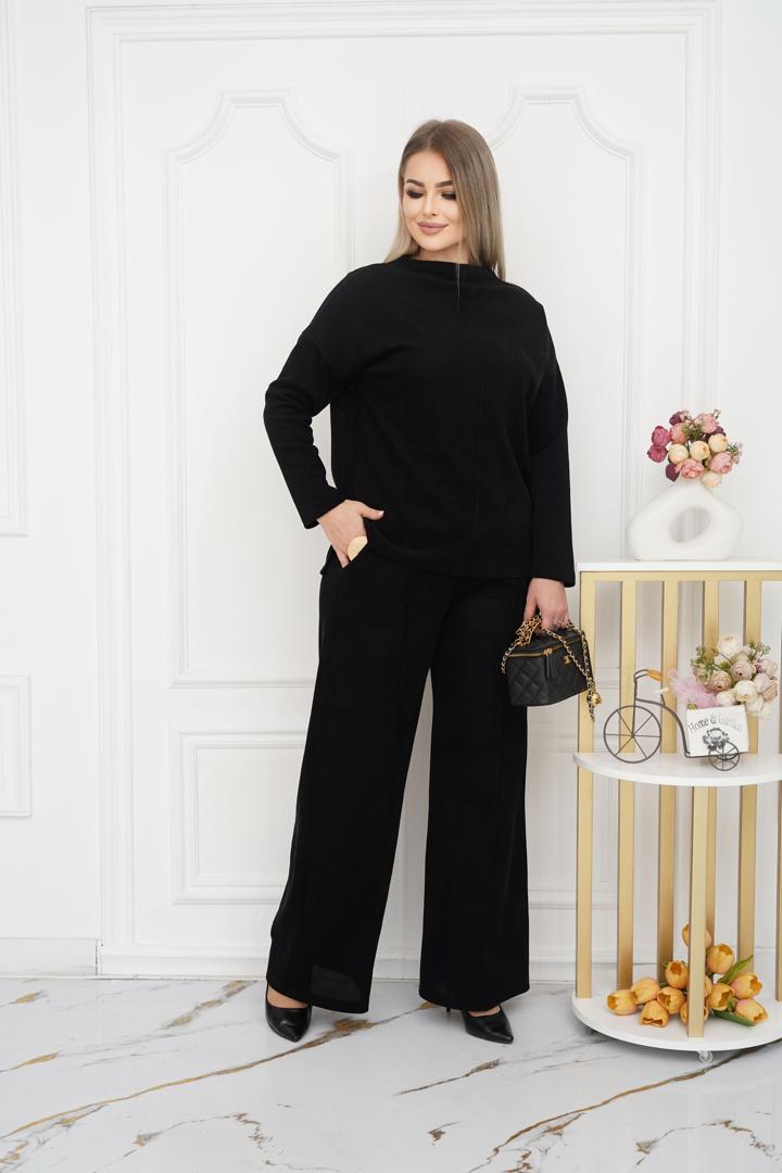 ქალის სარჩელი შავი თბილი დიდი ზომის - მოდელი აღებულა შავო ფერის კომბინირებული კოსტიუმით, რომელიც შედგება ფართო შარვლებიდან და ფართო მაისურიდან, ფონზე თეთრი კედლები და დეკორფარეხიანი უჯრა.